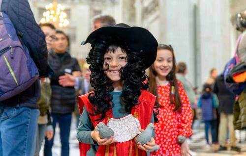 A young child wearing a long black, curly wig and wide brimmed hat, dressed as Christopher Wren.