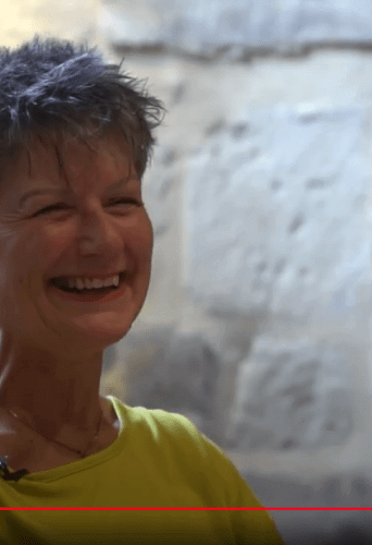 Claire Gilbert is a white woman with short grey hair wearing a yellow sweater and smiling to the audience. She stands in front of the sunlit alcove in the Wren Suite of the Cathedral.