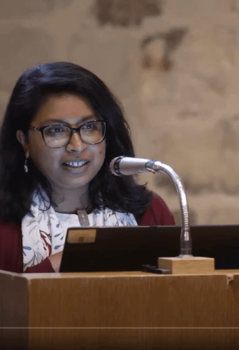 Anupama is a your Asian woman with shoulder length black hair wearing glasses. She is standing at a lectern with a microphone in the Wren Suite at St Paul's Cathedral.
