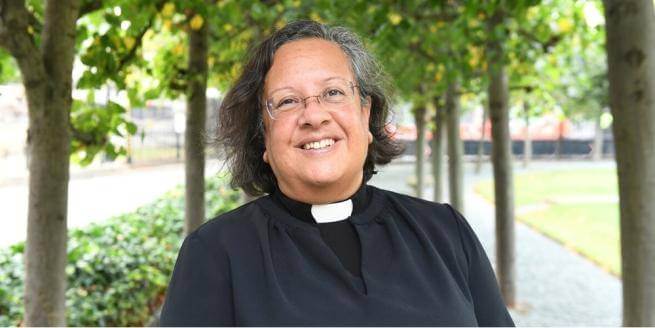 Tricia is pictured smiling standing outside under trees. She is a mixed-race woman with shoulder-length greying black hair and thin wire glasses, wearing a clerical collar under a black blouse.