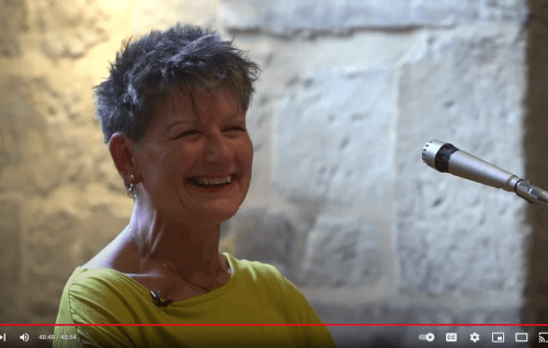 Claire Gilbert is a white woman with short grey hair wearing a yellow sweater and smiling to the audience. She stands in front of the sunlit alcove in the Wren Suite of the Cathedral.