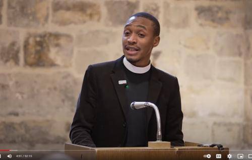 Jarel is a young black man wearing a clerical collar under a black shirt and suit, and he is standing at a lectern talking, in a room with an old exposed stone wall behind him.