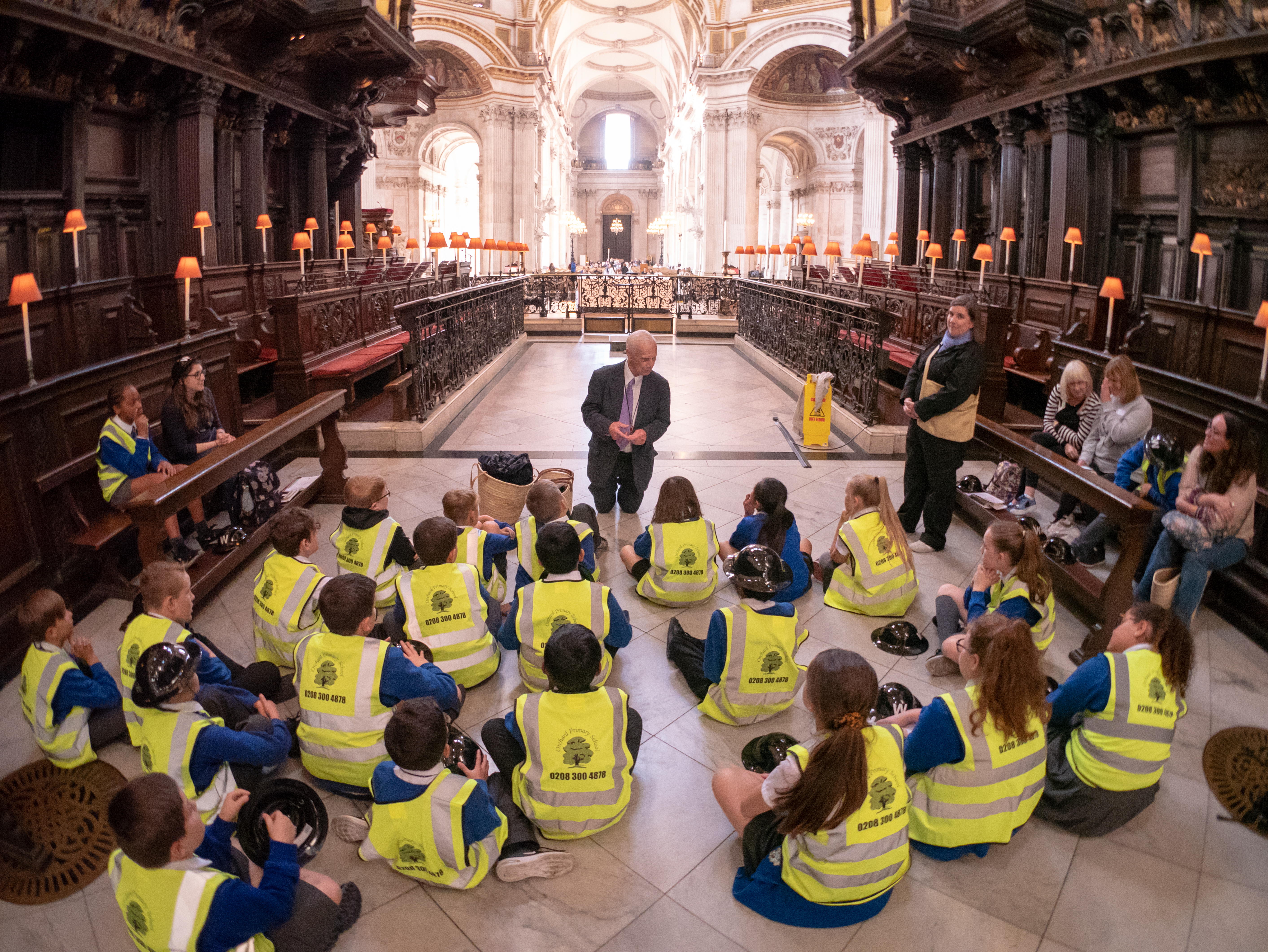 Children in blue uniforms and high vis lanyards sit on the floor of the quire listening to a talk from a cathedral volunteer