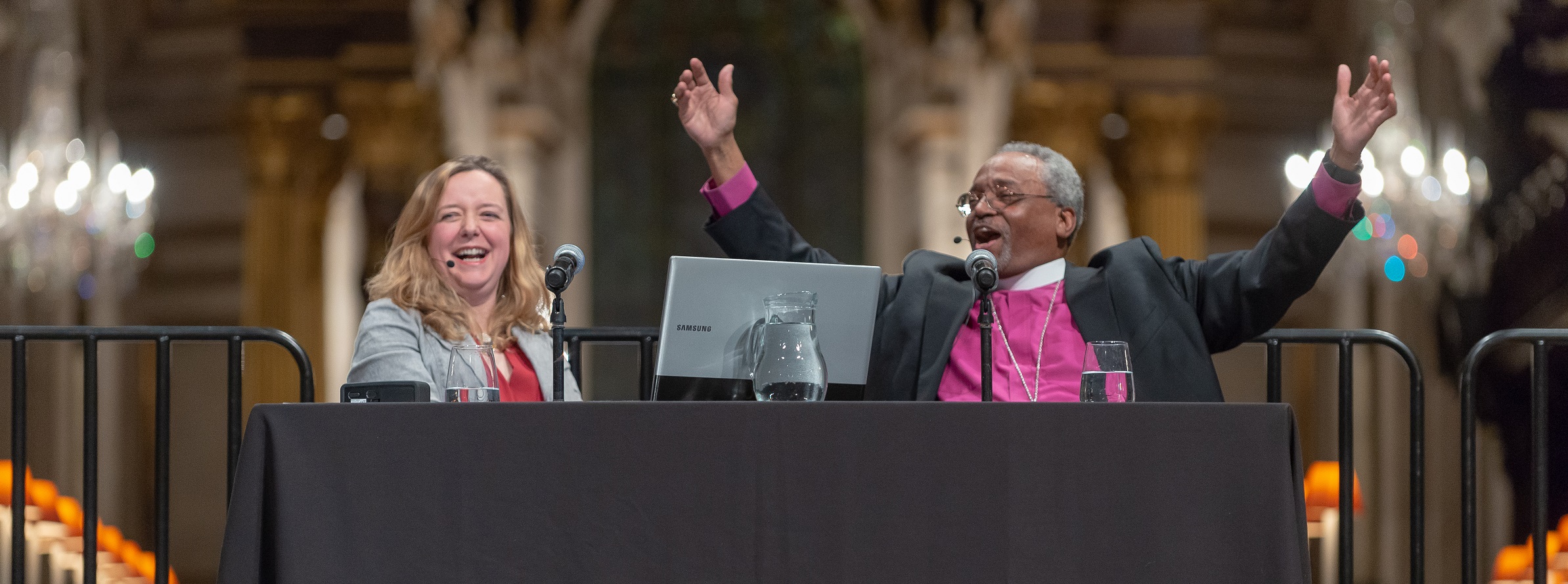 Bishop Curry, a black man with grey hair in a clerical collar purple shirt, on stage under the dome of the cathedral, smiling and arms outstretched, alongside a smiling Paula Gooder, a white woman with long blond hair in a grey jacket and red blouse.