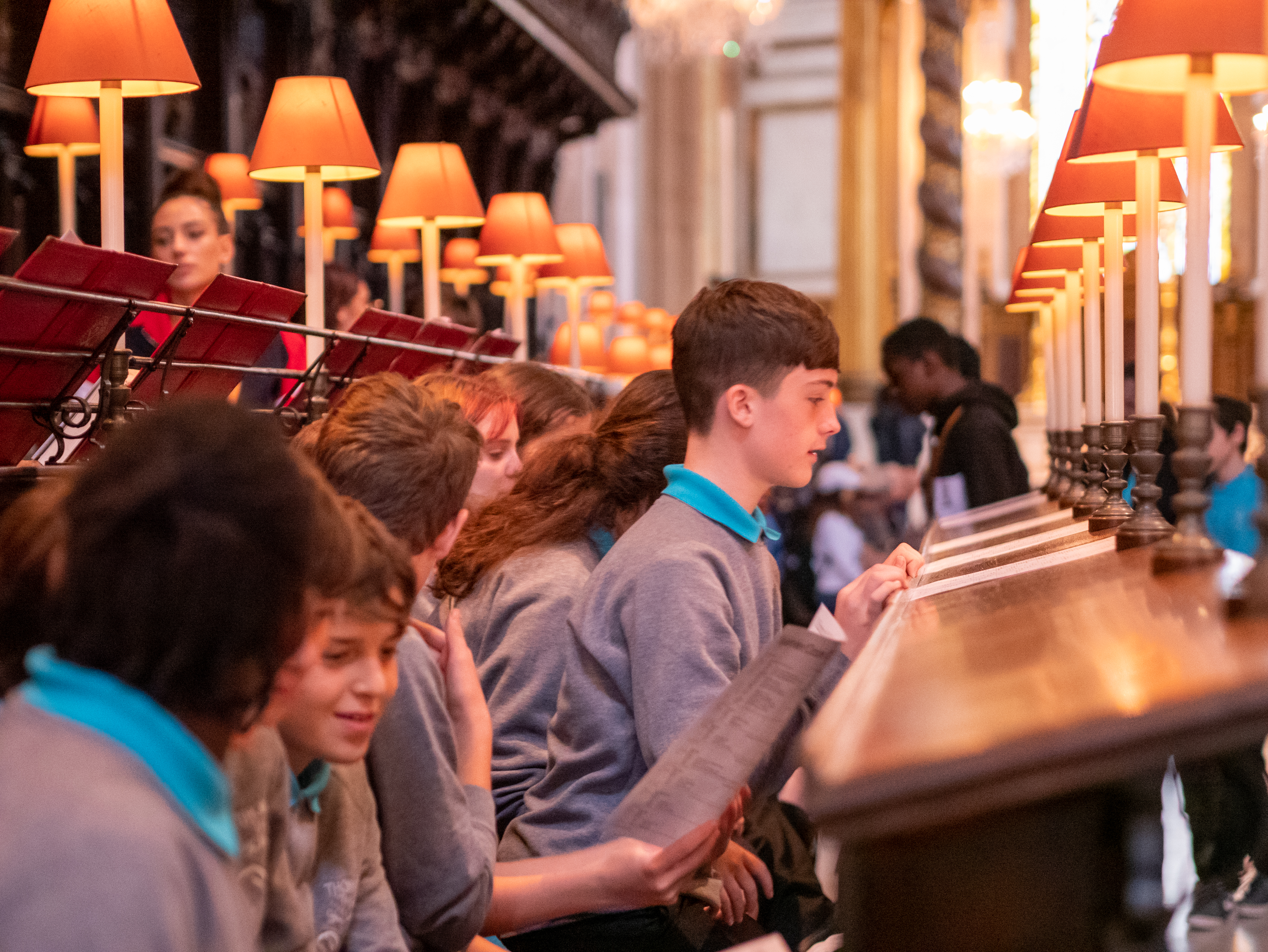 Secondary school age children in grey jumpers and blue polo shirts sit in the quire