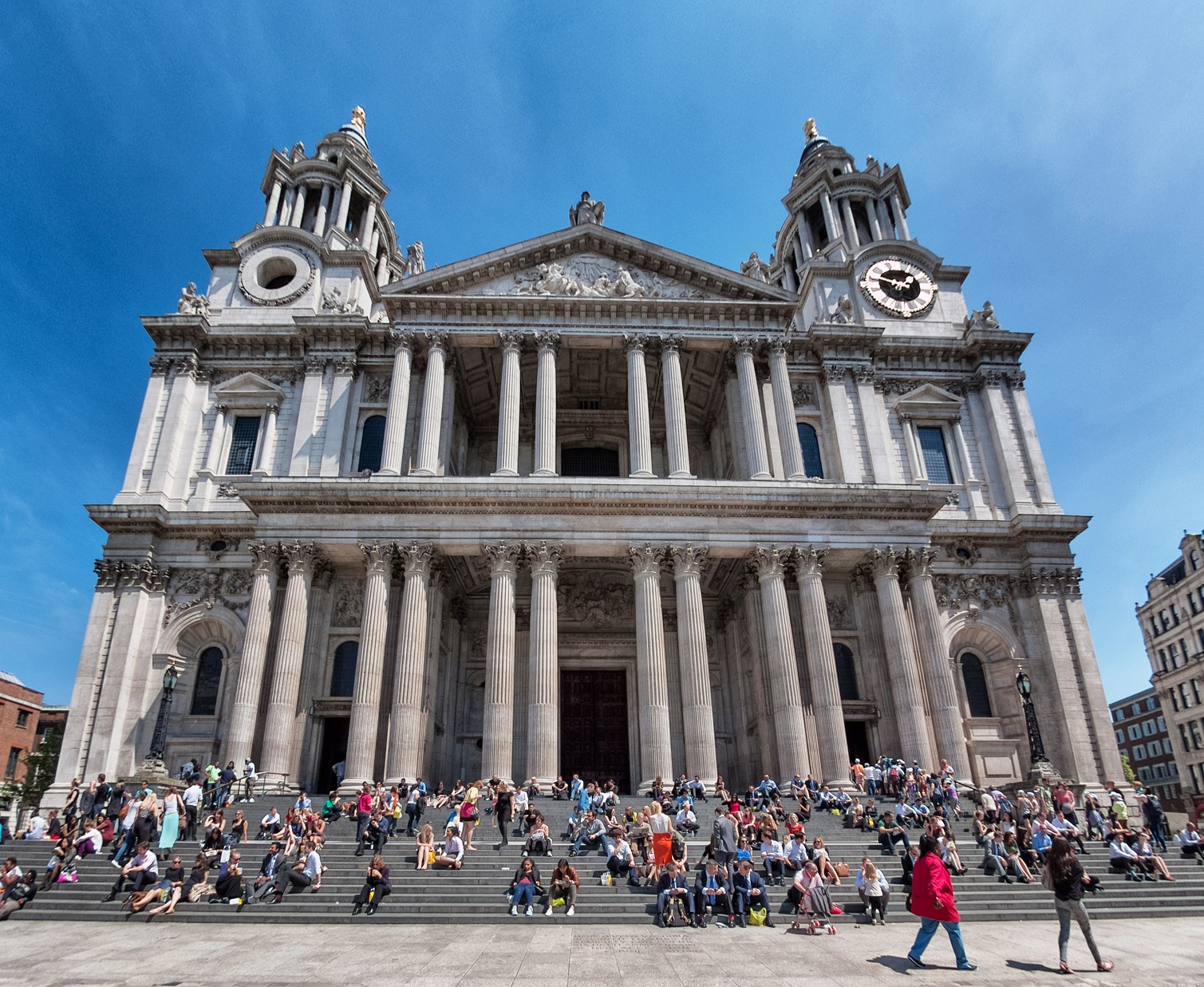The West front of the cathedral showing lots of people sitting on the steps up to the cathedral, with a sunny blue sky overhead