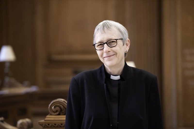 Alison has short grey hair and black-framed glasses. She wears a clerical collar and black shirt under a black jacket and stands by a wooden carving
