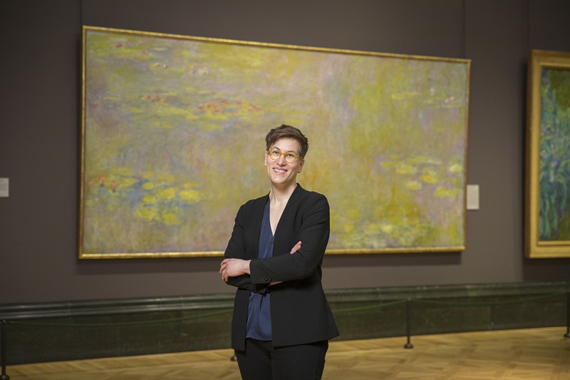 Ayla wears a black jacket and trousers with a dark blue blouse and has short brown hair and glasses, while standing in front of a painting in an art gallery