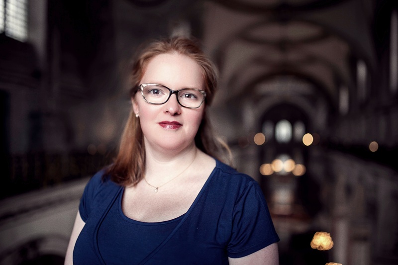 Carris wears a navy short sleeved dress and has long auburn hair and wears glasses. She stands at the triforium level of St Paul's Cathedral with lights glowing behind her