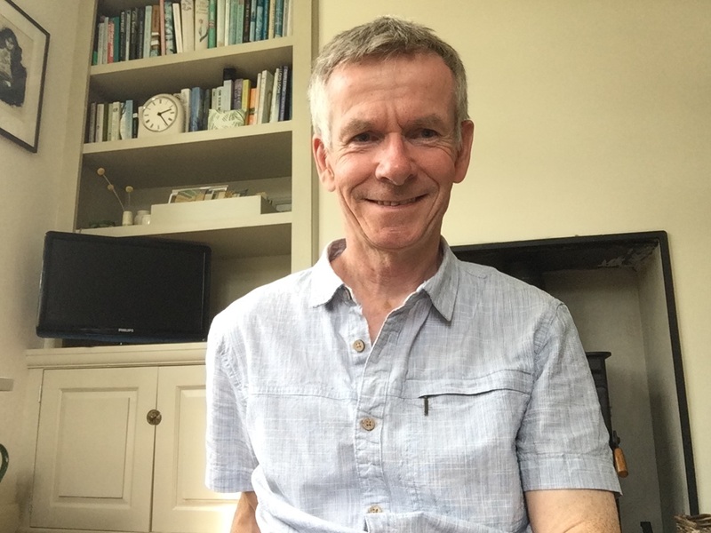 Christopher wears a blue linen short sleeved shirt and sits in a room with bookcase, tv and fireplace behind him