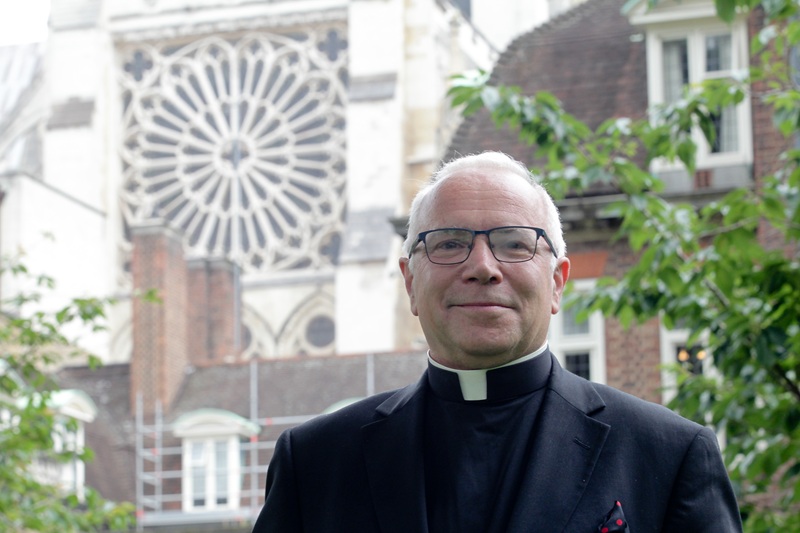 David wears a clerical collar with a black shirt and jacket, he has short white hair and glasses, and stands in a garden