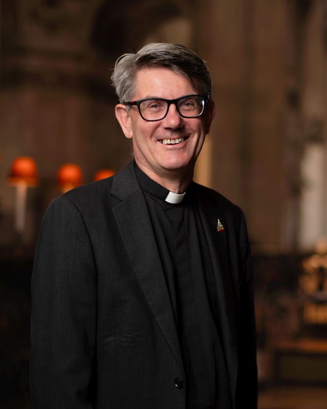 Andrew has short grey hair and wears glasses and a clerical collar and black shirt under a black jacket.