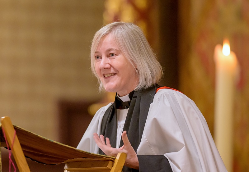 Ellen has a grey bob and is wearing a cassock and surplice, and stands at a lectern