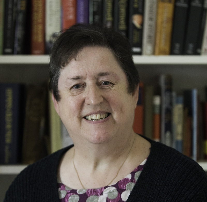 Gemma has short dark hair, wears a black cardigan over a patterned dress and stands in front of a full bookcase