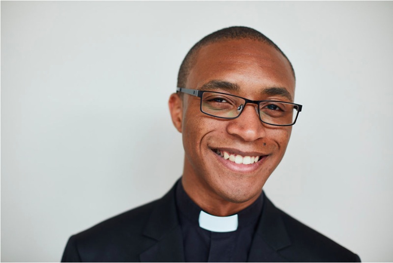 Jarel wears a clerical collar and black shirt, has shaved black hair and wears glasses.