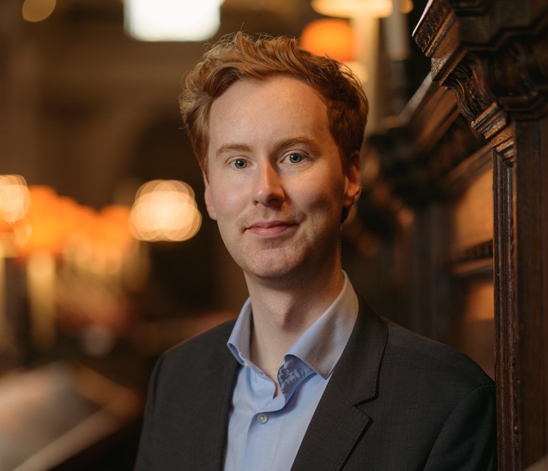 James has short auburn hair and wears a blue shirt under a dark grey jacket. He's in the quire of St Paul's with glowing lamps and carved woodworkbehind him.