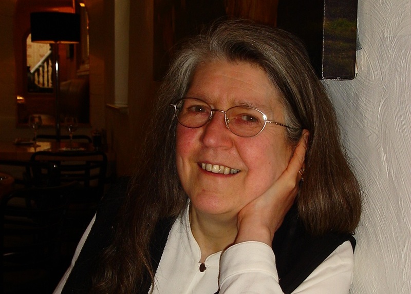 Janet wears a black waistcoat over a white blouse and has long dark hair, greying at the front, and glasses