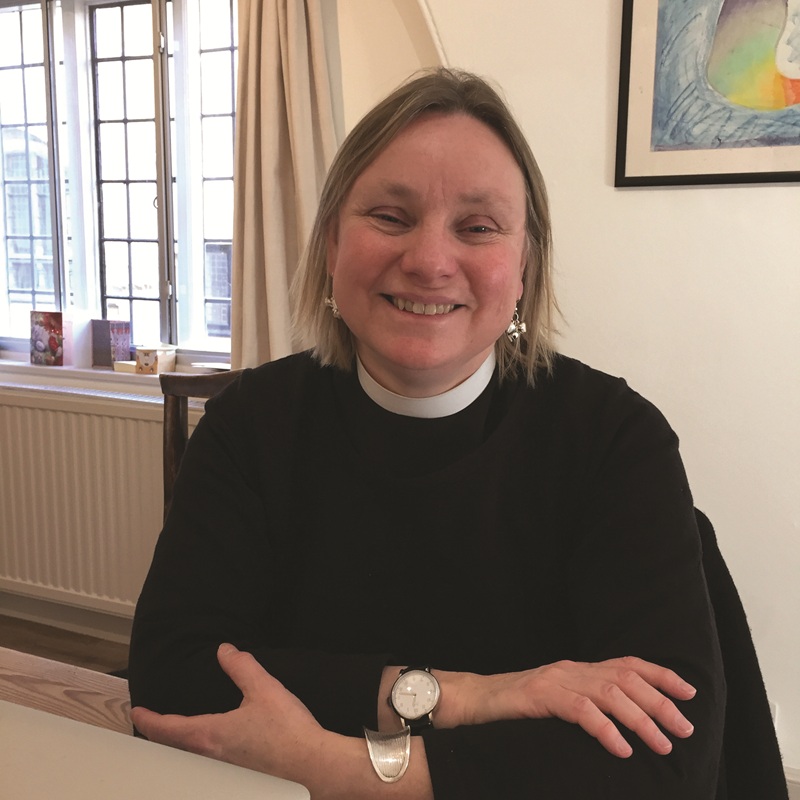 Jessica wears a clerical collar and black shirt, and has shortish fair hair. She ists on a dining chair in front of a panelled glass window.