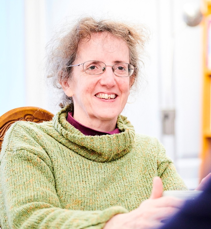 Karen has wavy light hair and wears glasses, she's wearing a pale green roll neck jumper and sitting on a wooden chair
