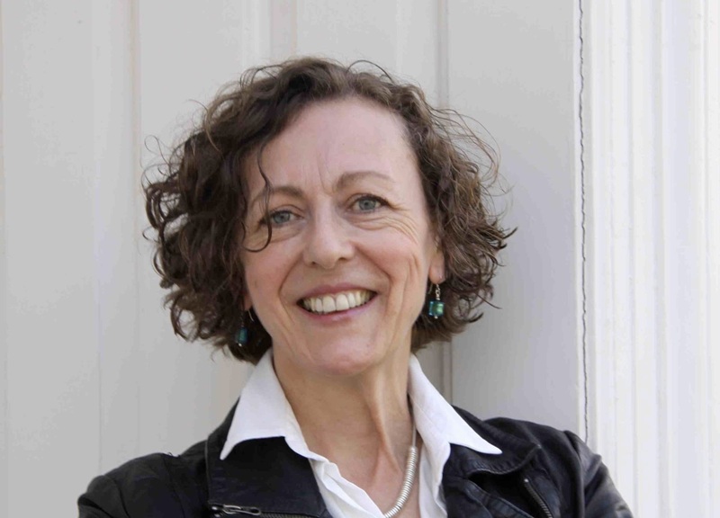 Maggi has short curly brown hair and wears a black jacket over a white shirt