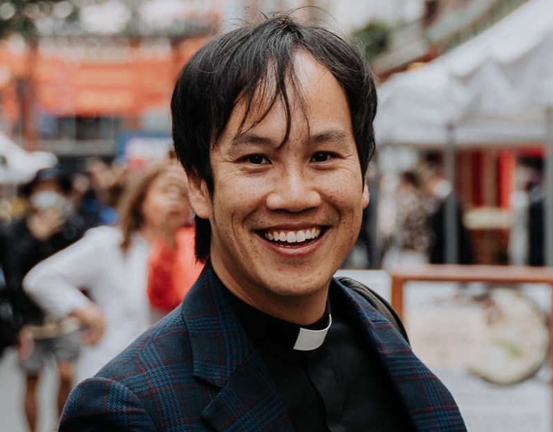 Mark has short dark brown hair and wears a clerical collar and black shirt under a checked jacket