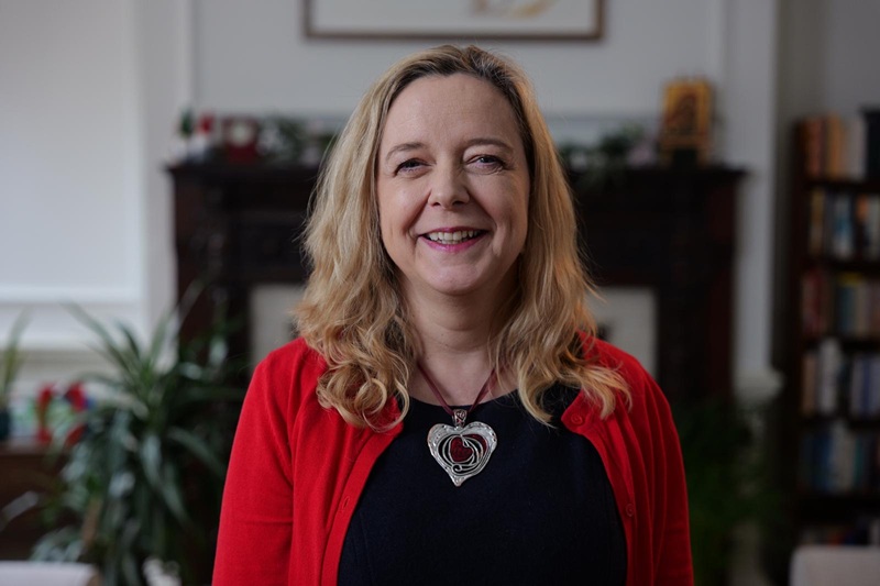 Paula has long blond hair and wears a red cardigan over a black top, standing in front of a fireplace