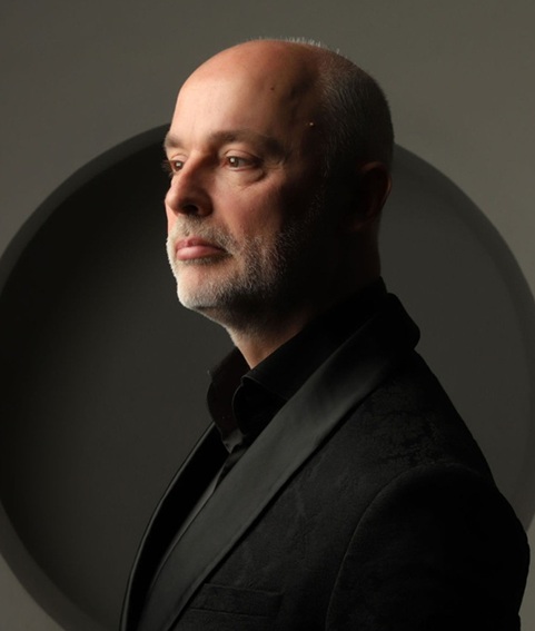 Patrick wears a black shirt under a black dinner jacket. He is balding and has a grey beard.