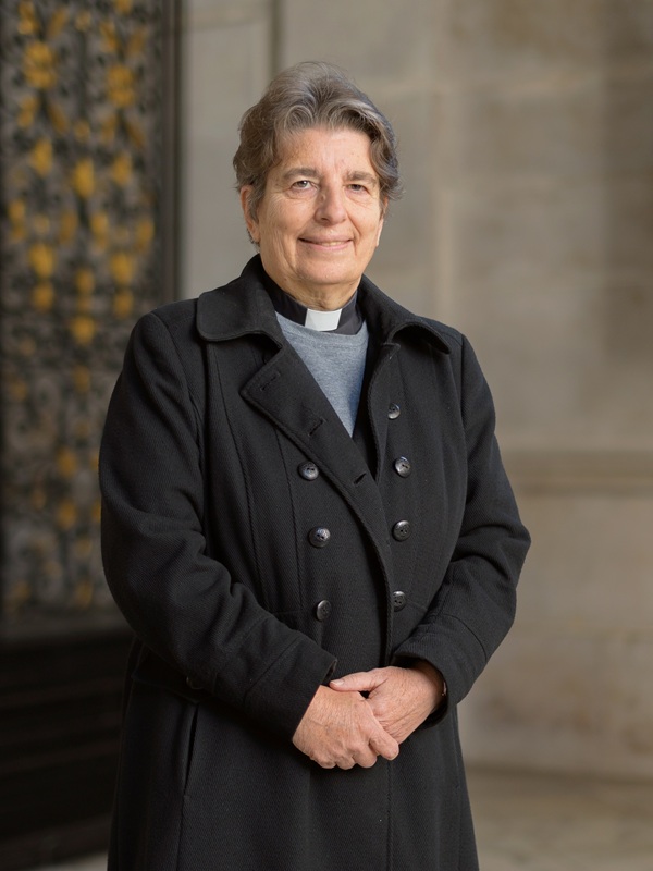 Paula wears a clerical collar under a grey jumper and a black overcoat.