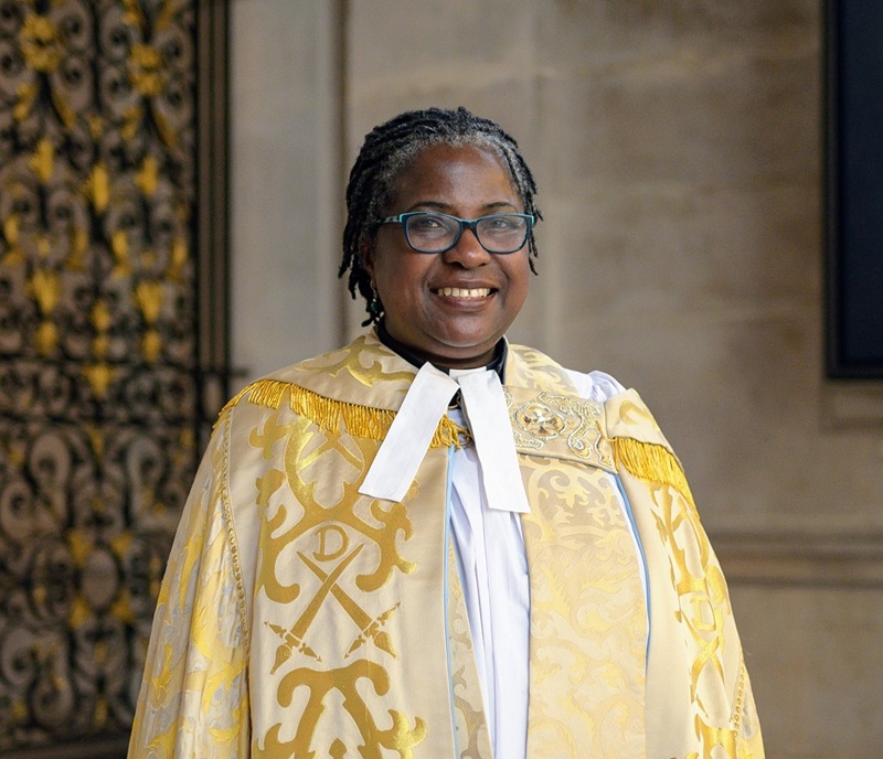 Ade has her hair in short dark braids and wears glasses, while wearing a golden robe over a white cassock and a preaching collar
