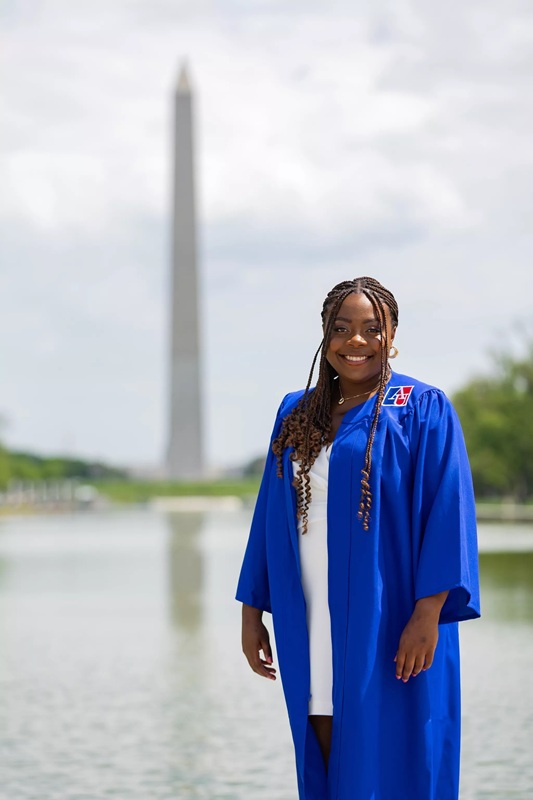 Alexandra has long dark hair in braids, wears a blue academic gown over a white dress and stands in front of the Washington monument alongside the lake