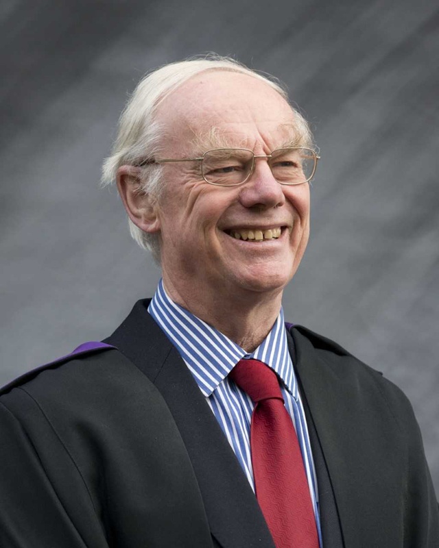 Ian wears a dark academic gown over a black suit, a red tie and a blue and white striped shirt. He has short white hair and wears glasses.
