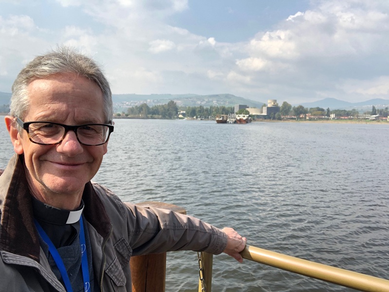 Richard, a man with short grey hair and glasses, wearing a clerical collar under a brown jacket, stands at the railing at the edge of a lake