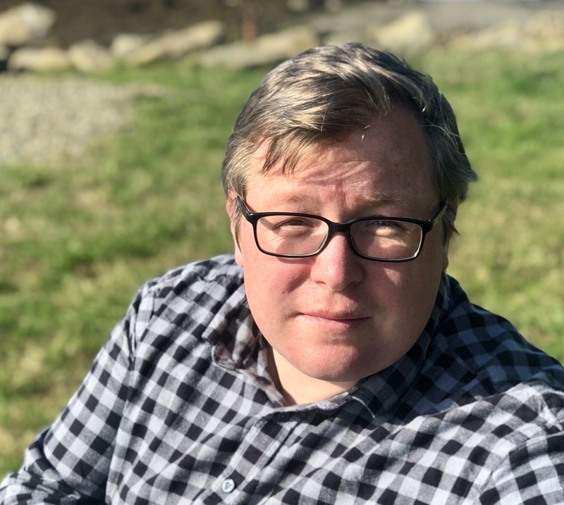 Sam has short blond hair and black glasses and wears a black and grey checked shirt