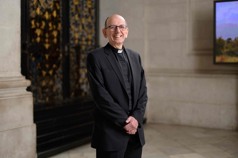 Neil wears a clerical collar under a black shirt and suite and stands in the Dean's Aisle at St Paul's Cathedral