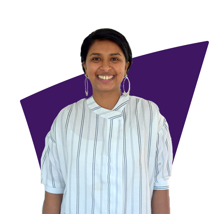 Anu has long dark hair worn in a ponytail and wears large silver drop earrings and a white shirt with grey stripes