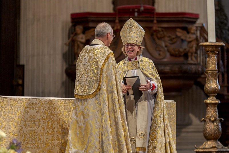 The then Bishop of London wearing a golden cope and mitre smiles at another member of clergy with his back to us, also wearing a golden cope, in the Cathedral