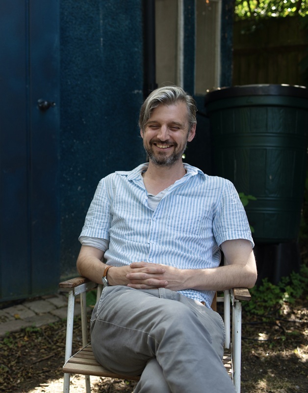 David has blond hair and a dark beard and wears a pale blue short sleeved striped shirt. He's sitting on a garden chair in the shade