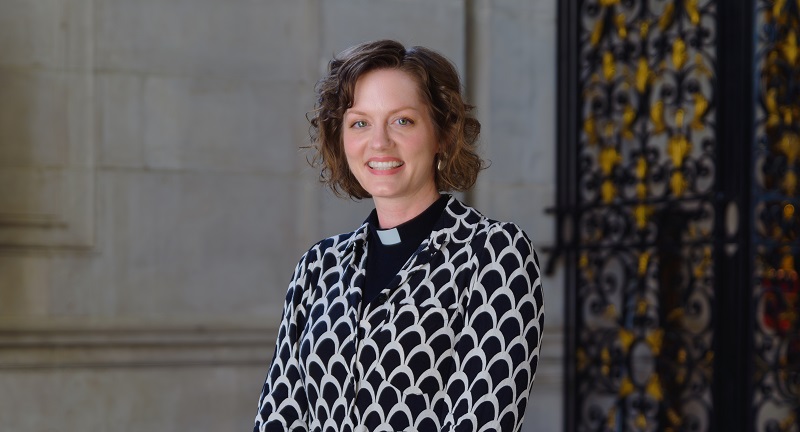 Tessa wears a clerical collar under a black and grey patterned dress and has shoulder length curly brown hair. She's standing by a decorated ironwork gate and a portland stone wall