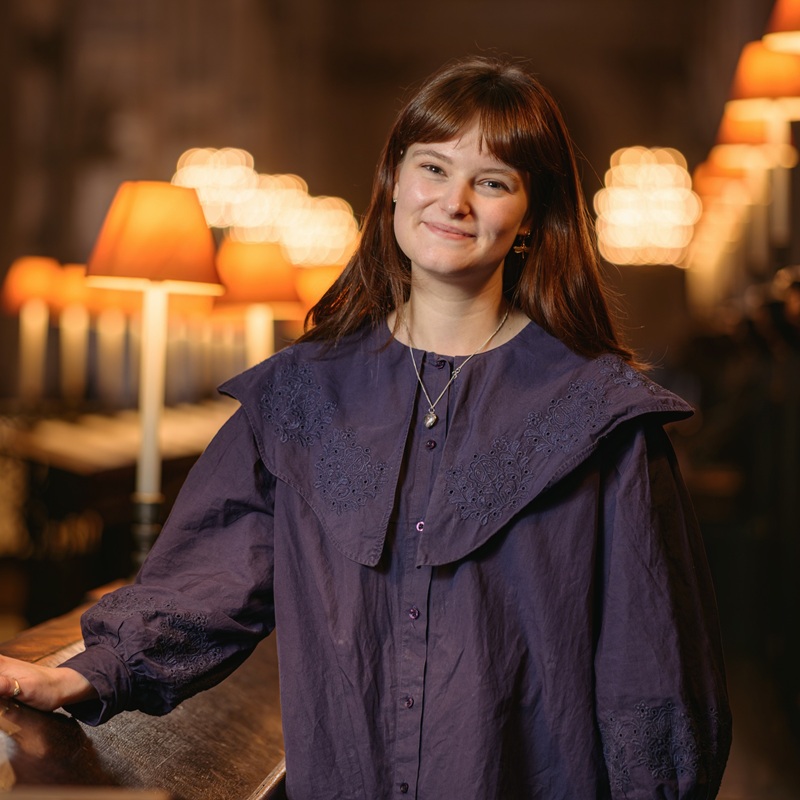 A young woman with long dark hair stands in the wooden stalls of the quire, lamps glowing in the background