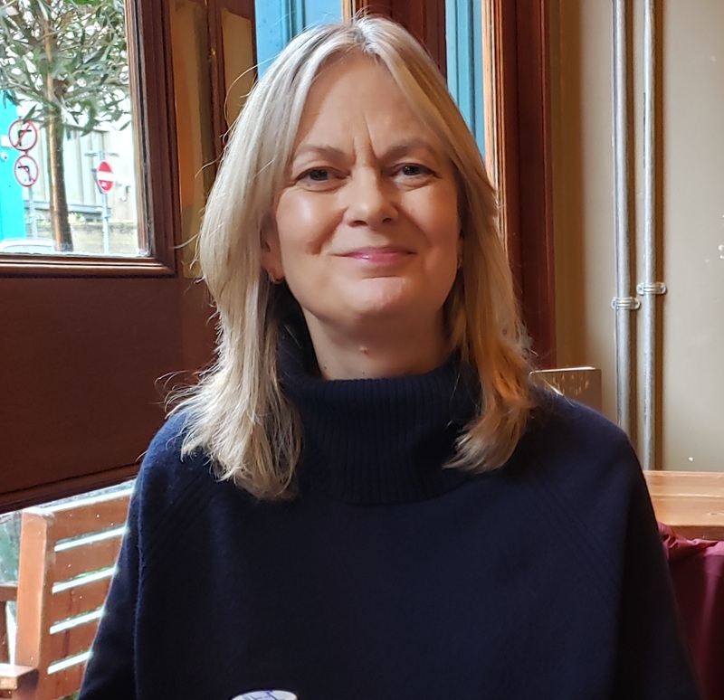Linda has long straight blond hair and wears a navy turtleneck jumper