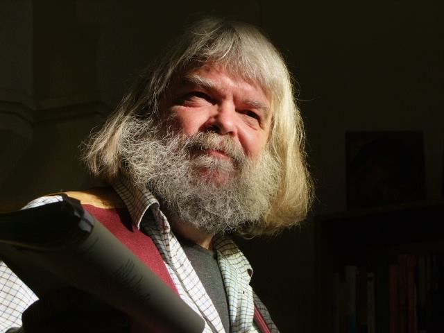 Malcolm has long grey hair and a grey beard, and wears an open necked checked shirt over a grey tshirt