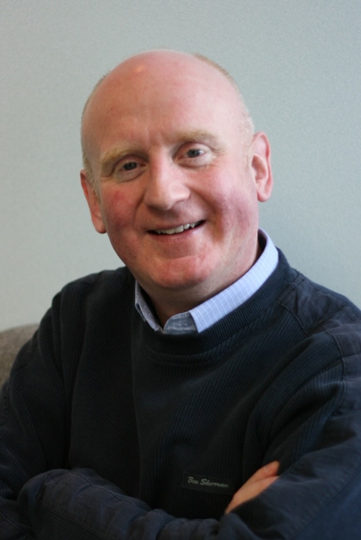 Peter has a bald head, wears a navy sweater over a pale blue shirt