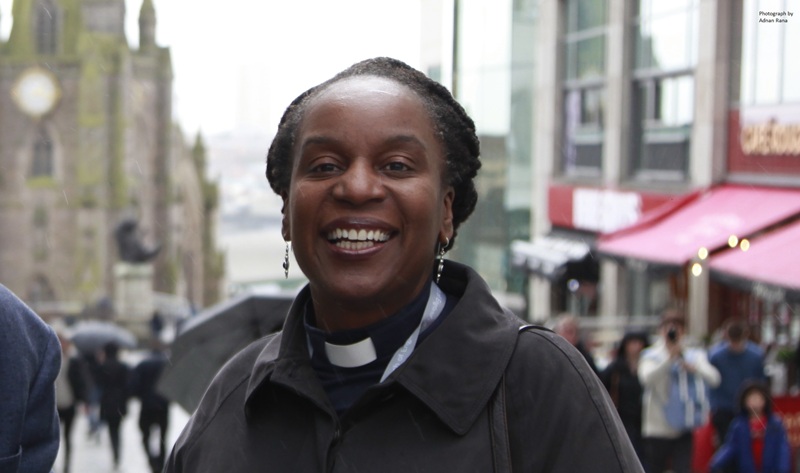 Sharon has short black hair, wears a clerical collar under a black jacket, and is photographed in a busy town centre
