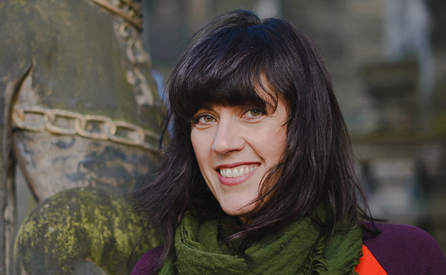 Debbie has long dark hair with a fringe, and wears a dark green scarf over a purple coat