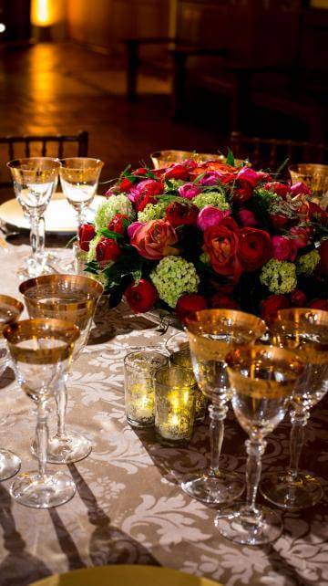 Pink, red and pale yellow flower arrangements on a white table cloth with glasses and cutlery set for dinner