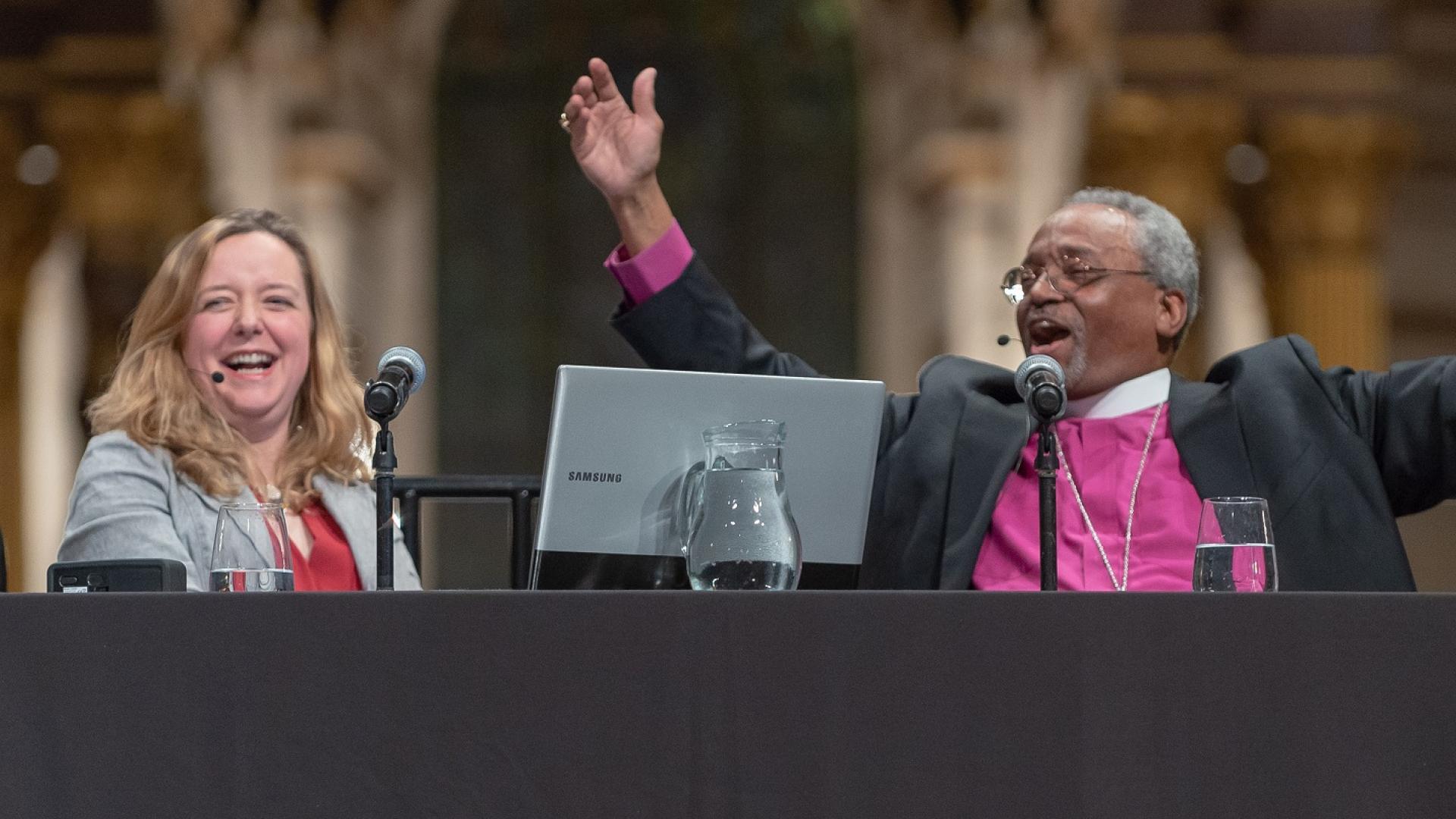 Bishop Curry raises his arms and Paula Gooder laughs alongside on the stage at an event at St Paul's Cathedral