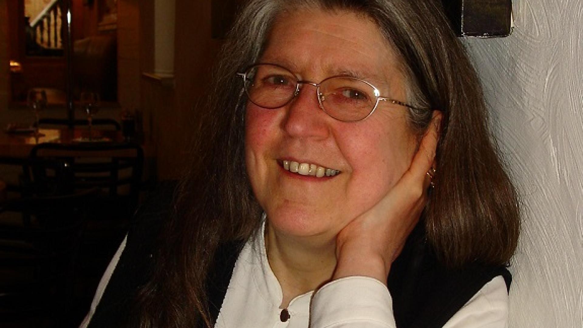 Janet Tollington leans her elbow on a table and with her hand under her chin smiles towards the camera