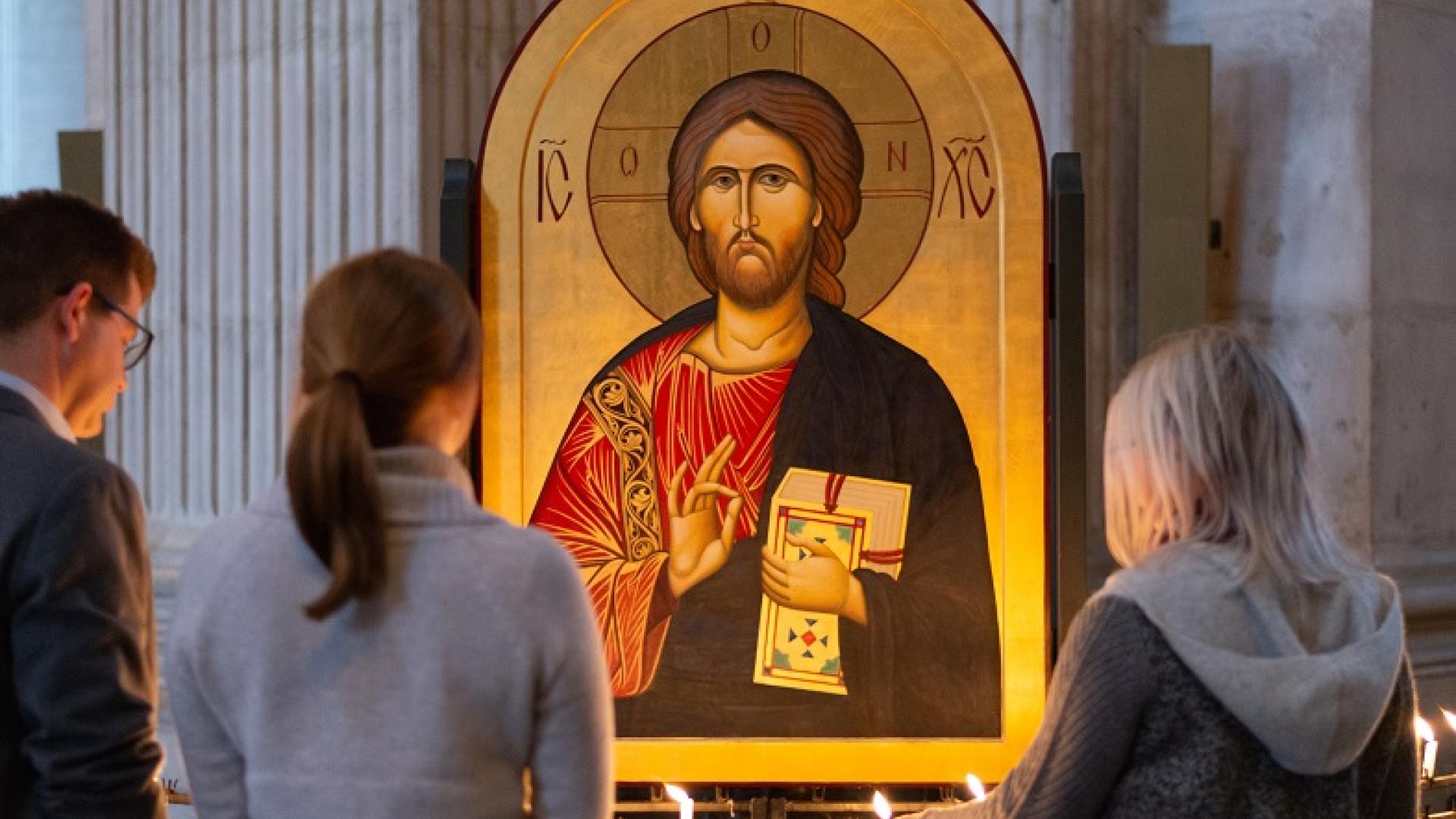 Two women and a man stand before an Icon of Jesus and light candles as they reflect.