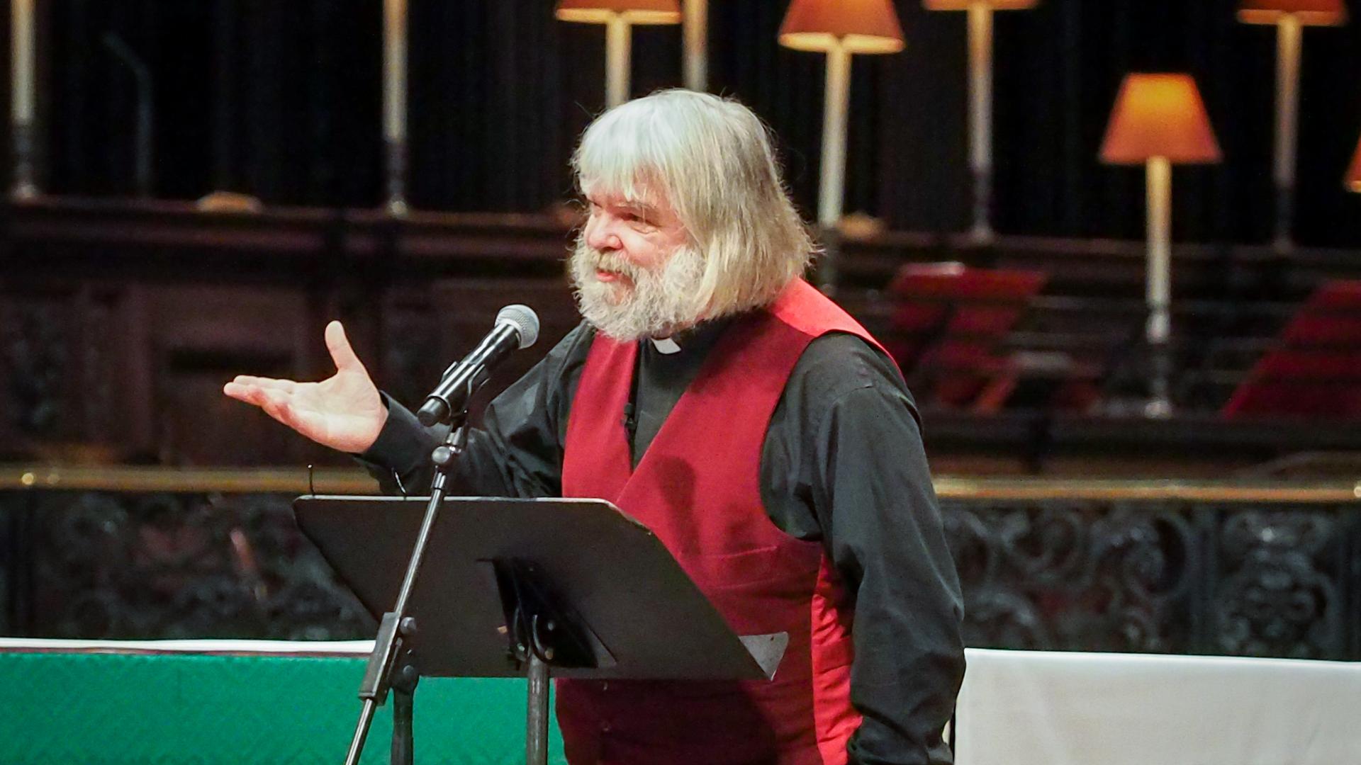 Malcolm Guite stands on the dais at St Paul's Cathedral, hand raised as he speaks, green altar cloth on the altar behind him.