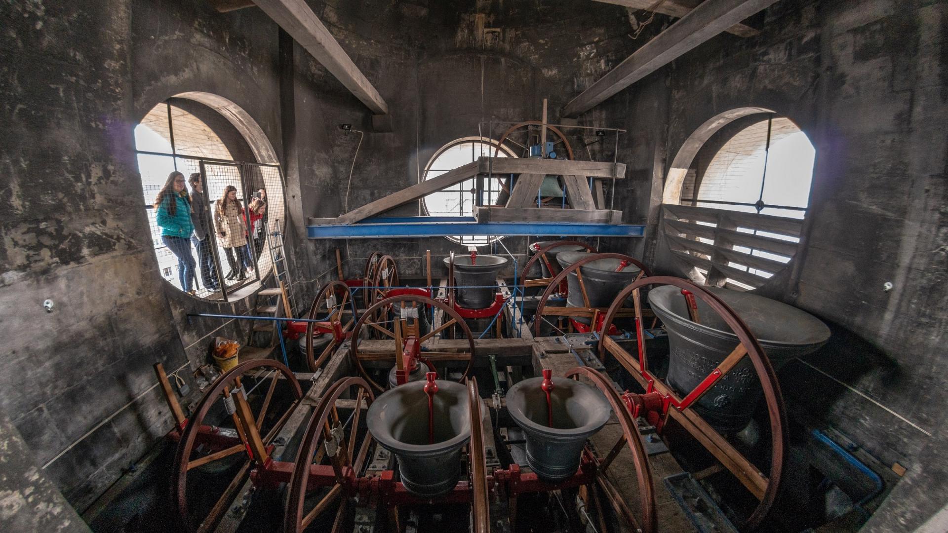 A wide angled shot from inside the north west tower, showing the 12 change ringing bells as they are ringing. Some children stand to one side outside the tower, on the roof, looking in.
